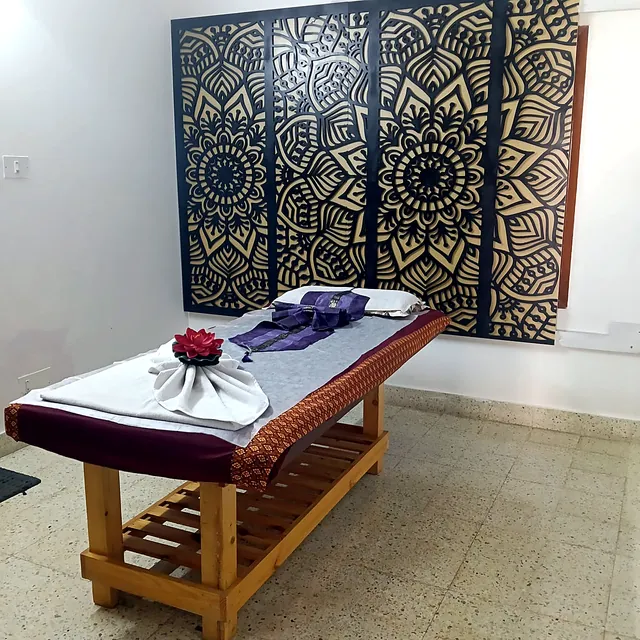 Treatment room at Le Grand Thai Spa with a wooden massage bed, Thai silk runner, folded towels and a carved mandala wall panel